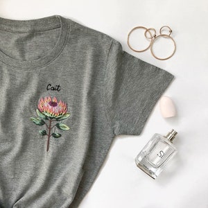 May include: Gray t-shirt with a pink and yellow embroidered flower and the word "Cait" above it.
