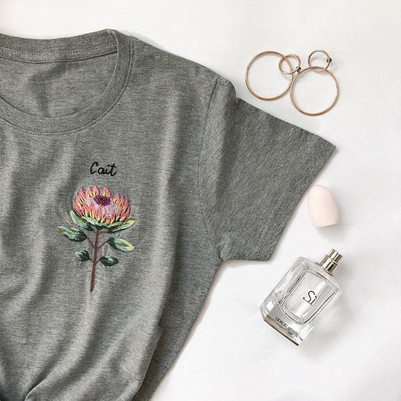 May include: Gray t-shirt with a pink and yellow embroidered flower and the word "Cait" above it.