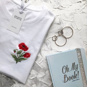 May include: White t-shirt with embroidered red poppies and a white tag that says "Vyshyto". The t-shirt is lying on a white textured surface next to a blue notebook with the text "Oh My Book! Mother edition" and gold hoop earrings.