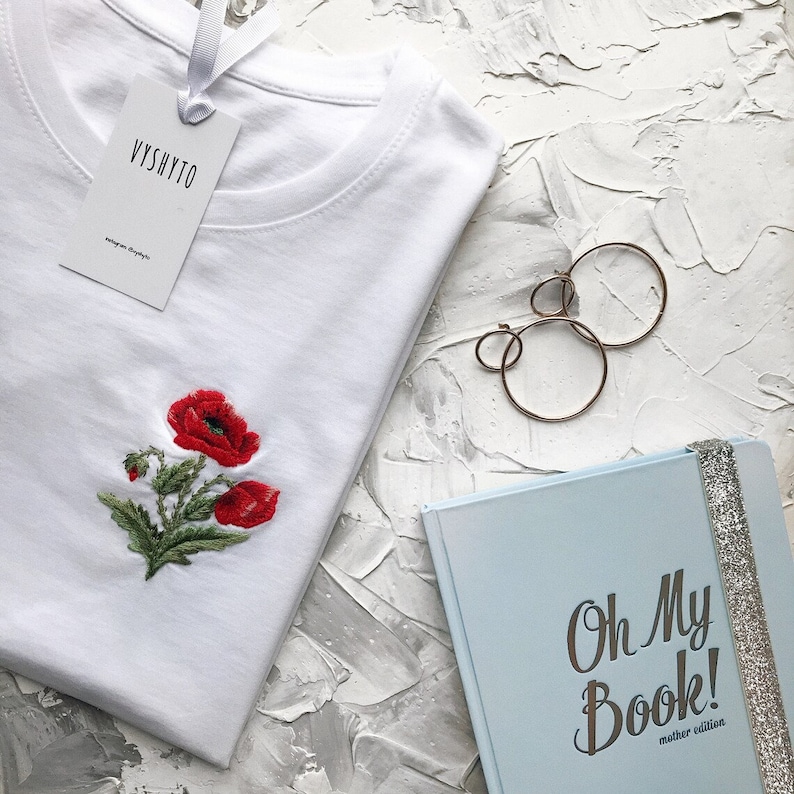 May include: White t-shirt with embroidered red poppies and a white tag that says "Vyshyto". The t-shirt is lying on a white textured surface next to a blue notebook with the text "Oh My Book! Mother edition" and gold hoop earrings.
