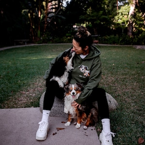 May include: A woman wearing a green hoodie with a dog print sits on a rock with two dogs. The hoodie has the text "KIXX + CATT" on it. The woman is wearing white trainers and white socks.