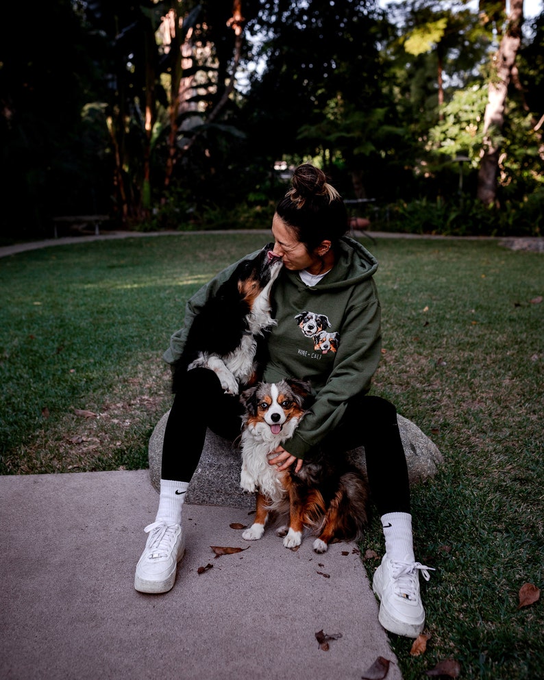 May include: A woman wearing a green hoodie with a dog print sits on a rock with two dogs. The hoodie has the text "KIXX + CATT" on it. The woman is wearing white trainers and white socks.