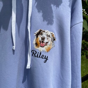 May include: A light blue hoodie with a white drawstring. The hoodie features an embroidered image of a dog's head with the name "Riley" underneath.