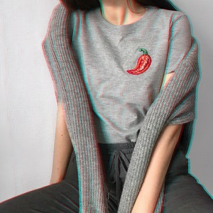 May include: A grey t-shirt with a red chilli pepper embroidered on the front. The shirt is worn with a grey cardigan and black trousers.