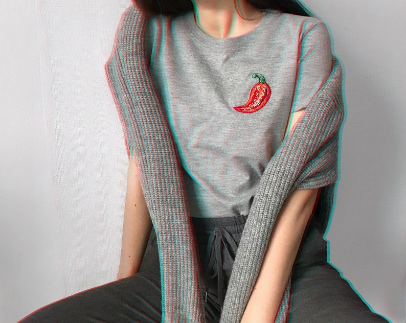 May include: A grey t-shirt with a red chilli pepper embroidered on the front. The shirt is worn with a grey cardigan and black trousers.