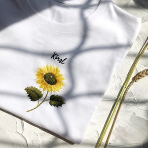 May include: White t-shirt with an embroidered sunflower and the name "Kirst" in black script.