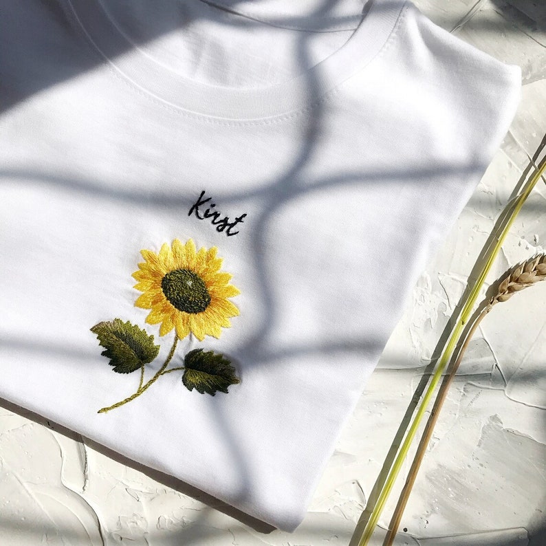 May include: White t-shirt with an embroidered sunflower and the name "Kirst" in black script.