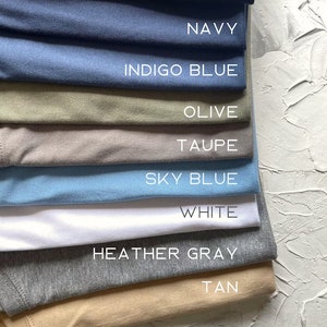 May include: A stack of t-shirts in various colors, including black, navy, indigo blue, olive, taupe, sky blue, white, heather gray, and tan.