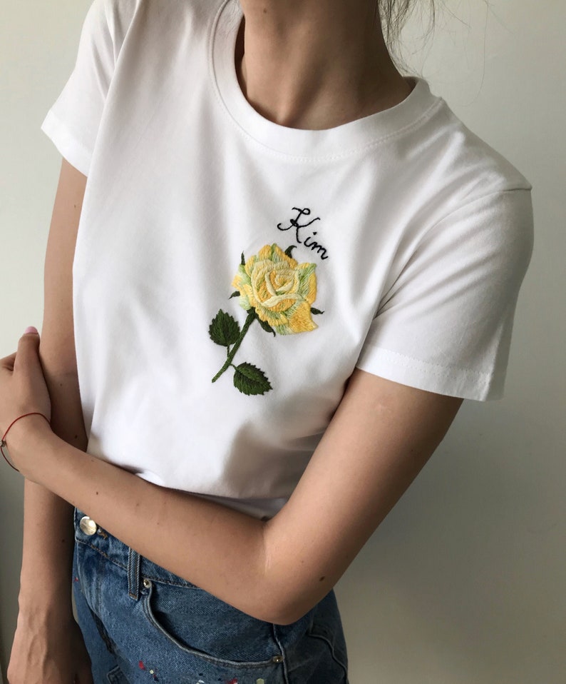 May include: White t-shirt with a yellow embroidered rose and the word "Kim" in cursive.