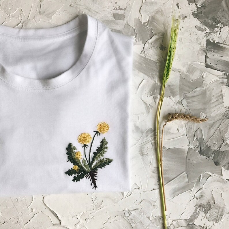 May include: White t-shirt with a yellow and green embroidered dandelion design.