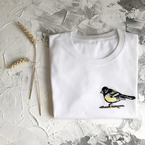 May include: White t-shirt with an embroidered black and yellow bird on a branch.