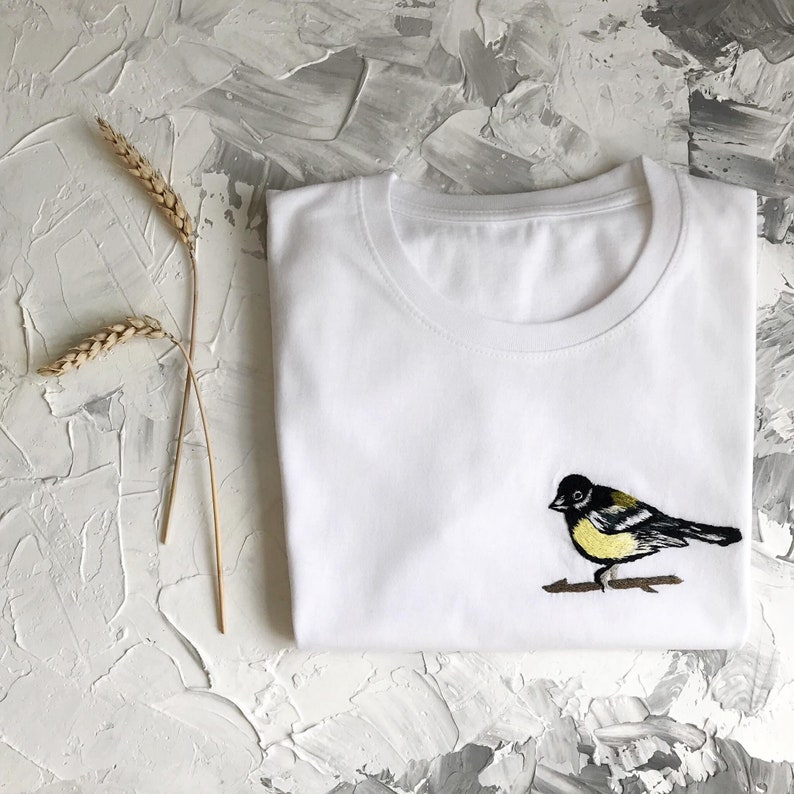 May include: White t-shirt with an embroidered black and yellow bird on a branch.