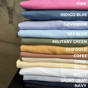 May include: A stack of 14 t-shirts in various colours, including pink, indigo blue, lavender, sky blue, military green, old gold, coffee, sand, beige, white, sport grey, navy, and black.