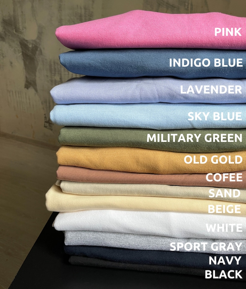 May include: A stack of 14 t-shirts in various colours, including pink, indigo blue, lavender, sky blue, military green, old gold, coffee, sand, beige, white, sport grey, navy, and black.
