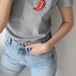 May include: A grey t-shirt with a red embroidered chilli pepper on the front. The shirt is paired with light blue jeans.