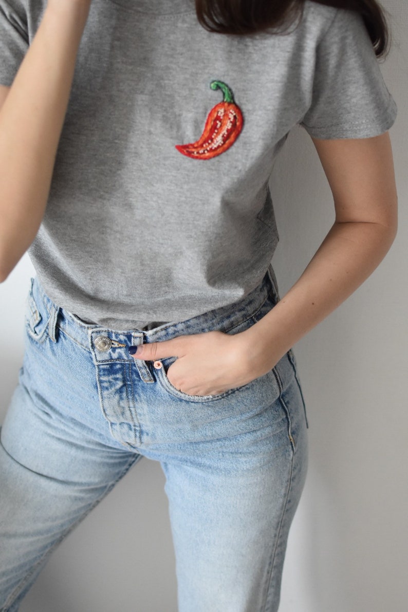 May include: A grey t-shirt with a red embroidered chilli pepper on the front. The shirt is paired with light blue jeans.