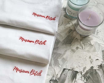 May include: Three white t-shirts stacked on top of each other. Each shirt has the phrase "Museum Bitch" embroidered in red on the front.