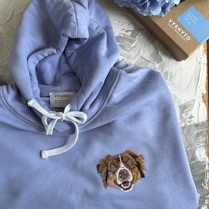 May include: A light blue hoodie with a white drawstring and an embroidered image of a brown and white dog. The hoodie has the text "VYSHYTO" and "BE THE PERSON WHO DOG THINKS YOU ARE" on a white tag.