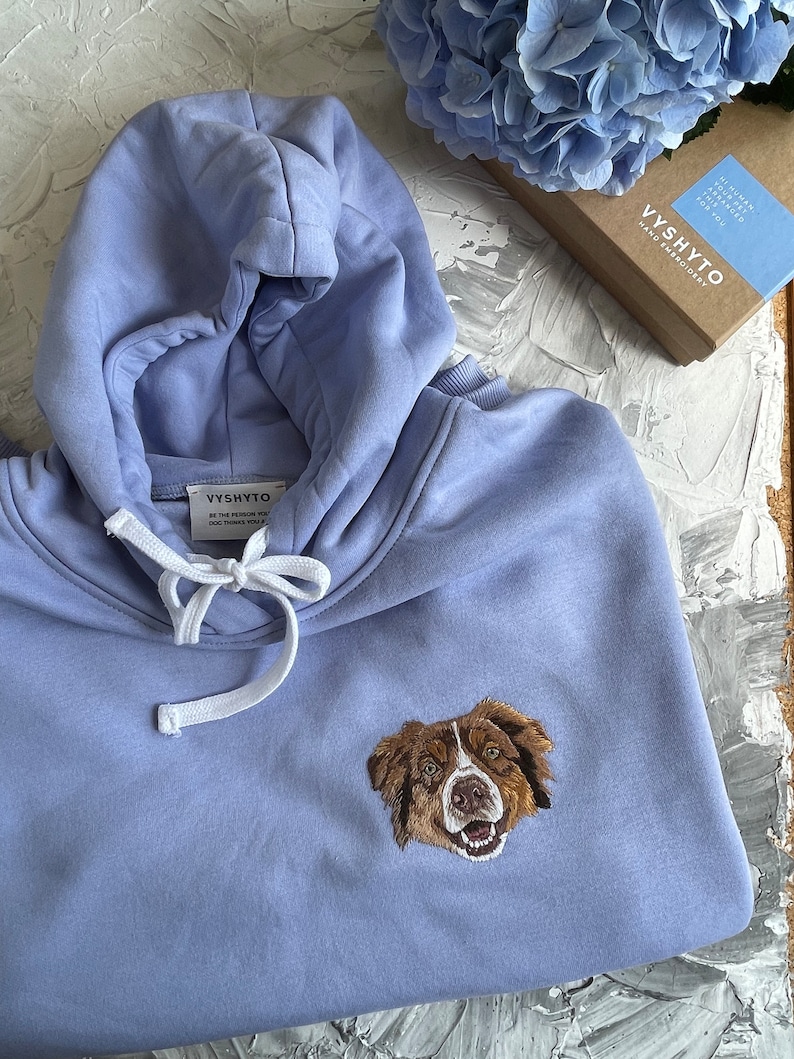May include: A light blue hoodie with a white drawstring and an embroidered image of a brown and white dog. The hoodie has the text "VYSHYTO" and "BE THE PERSON WHO DOG THINKS YOU ARE" on a white tag.