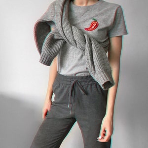 May include: A grey t-shirt with a red chilli pepper embroidered on the front. The t-shirt is worn with a grey knitted scarf and grey sweatpants.