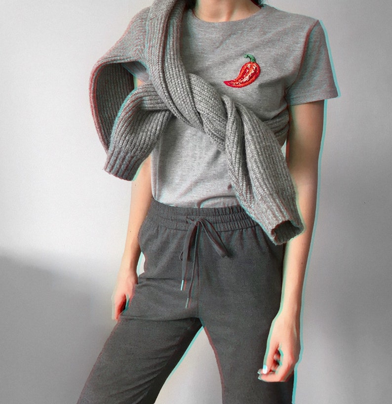 May include: A grey t-shirt with a red chilli pepper embroidered on the front. The t-shirt is worn with a grey knitted scarf and grey sweatpants.