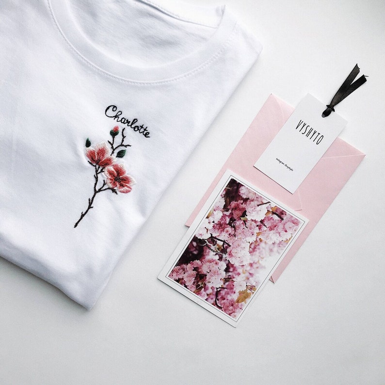 May include: White t-shirt with the name "Charlotte" embroidered on the front. The shirt features a small embroidered flower with pink and red petals. The t-shirt is displayed with a pink envelope and a card with a floral design.