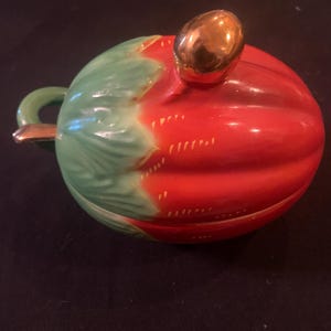 May include: A ceramic, lidded container with a red body and green top, resembling a tomato. The lid has a gold-colored knob and the handle is also gold. The container is likely for serving or storage.