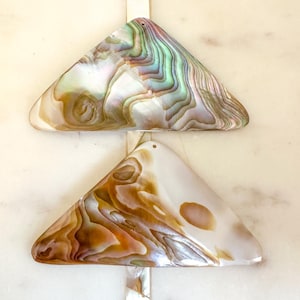 Puede incluir: Dos colgantes triangulares de concha iridiscente con un agujero en la parte superior para colgar. El colgante superior tiene un patrón iridiscente verde y azul, mientras que el colgante inferior tiene un patrón iridiscente marrón y blanco.