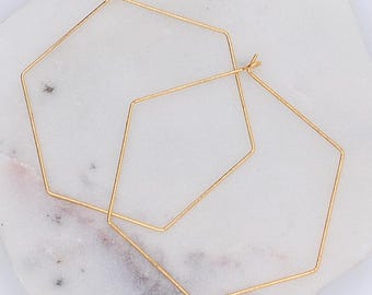 Puede incluir: Dos pendientes de aro geométricos de color dorado. Cada pendiente tiene forma de hexágono de alambre. Los pendientes se muestran sobre una superficie de mármol blanco y gris. Los pendientes son sencillos y modernos.
