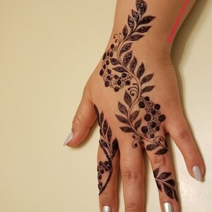 Puede incluir: Un primer plano de una mano con intrincados dise&ntilde;os de henna en forma de hojas y puntos. La henna es de color marr&oacute;n oscuro y cubre el dorso de la mano y los dedos. Las u&ntilde;as est&aacute;n pintadas de un color plateado claro.