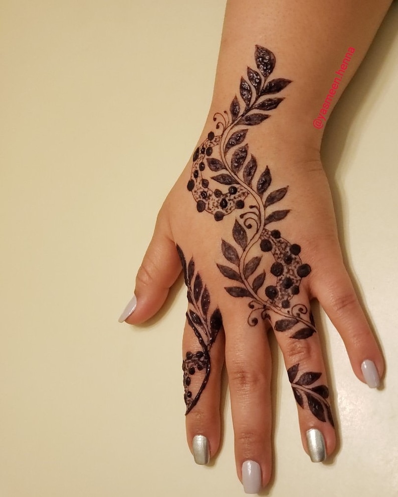 Puede incluir: Un primer plano de una mano con intrincados dise&ntilde;os de henna en forma de hojas y puntos. La henna es de color marr&oacute;n oscuro y cubre el dorso de la mano y los dedos. Las u&ntilde;as est&aacute;n pintadas de un color plateado claro.