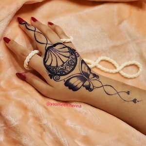 Puede incluir: Un dise&ntilde;o de tatuaje de henna negro en una mano con un motivo de mariposa y concha. El tatuaje est&aacute; en la mano de una persona e incluye el texto "@yasmeen.henna".