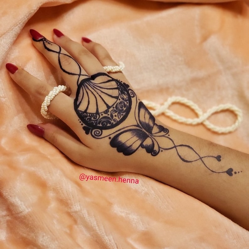 Puede incluir: Un dise&ntilde;o de tatuaje de henna negro en una mano con un motivo de mariposa y concha. El tatuaje est&aacute; en la mano de una persona e incluye el texto "@yasmeen.henna".