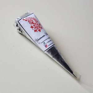 Puede incluir: Un cono de henna negro con una etiqueta blanca que dice "Yasmeen Henna" e incluye informaci&oacute;n de contacto. El cono est&aacute; sobre una superficie blanca.