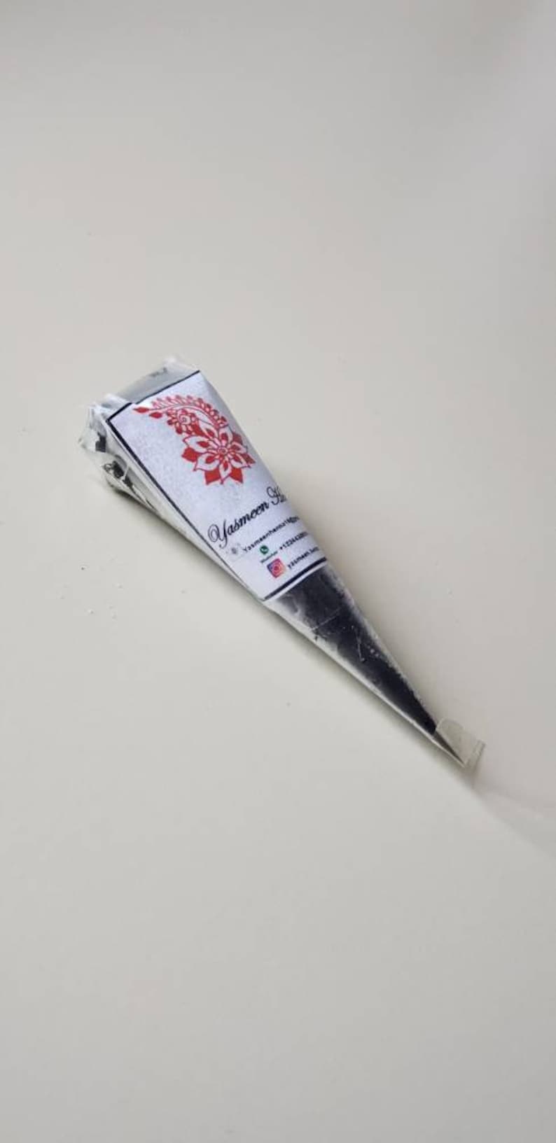 Puede incluir: Un cono de henna negro con una etiqueta blanca que dice "Yasmeen Henna" e incluye informaci&oacute;n de contacto. El cono est&aacute; sobre una superficie blanca.