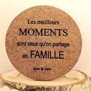 Puede incluir: Un posavasos redondo de corcho con el texto "Les meilleurs MOMENTS sont ceux qu'on partage en FAMILLE" en letras negras. El posavasos tiene un diseño decorativo de flechas y un corazón.