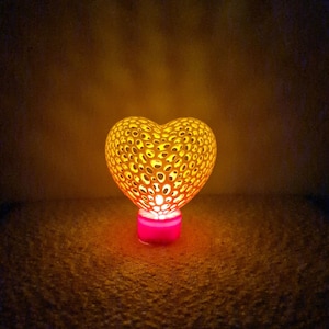 Può includere: Una lampada a forma di cuore con un design delicato e intricato. La lampada è realizzata in un materiale translucido ed emette una luce calda e gialla. La base della lampada è un piccolo cilindro rosa.