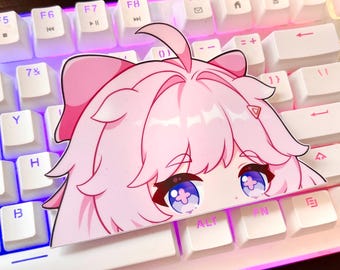 Puede incluir: Una pegatina de personaje de anime rosa con ojos grandes y un lazo rosa en la cabeza. La pegatina está colocada en un teclado blanco con iluminación arcoíris.