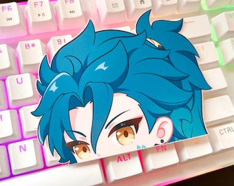May include: A sticker of a cartoon character with blue hair, a yellow eye, and a pink ear. The sticker is placed on a keyboard with white keys and pink accents.