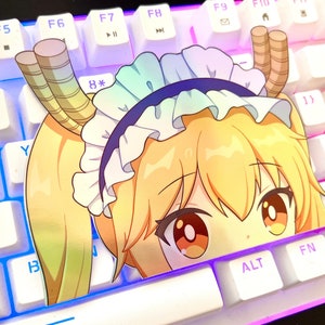 Può includere: Una tastiera bianca con illuminazione blu e viola. Uno sticker di un personaggio anime con capelli gialli, un vestito bianco e una fascia blu è sulla tastiera. Gli occhi del personaggio sono marroni e lo sticker copre parzialmente i tasti.