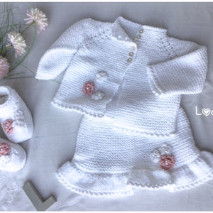 May include: White knitted baby outfit set with a cardigan, skirt, and booties. The cardigan and skirt have delicate white crocheted flowers with pink centers. The booties have pink flowers and pearl buttons.
