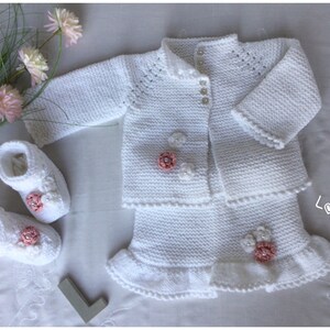 May include: White knitted baby outfit with a cardigan, skirt, and booties. The outfit is decorated with pink flowers and pearl buttons. The skirt has a ruffled hem. The booties have a pink flower detail. The outfit is laid out on a white surface with a bouquet of pink flowers.