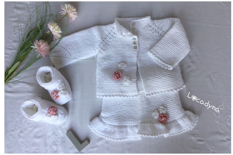 May include: White knitted baby outfit with a cardigan, skirt, and booties. The outfit is decorated with pink flowers and pearl buttons. The skirt has a ruffled hem. The booties have a pink flower detail. The outfit is laid out on a white surface with a bouquet of pink flowers.