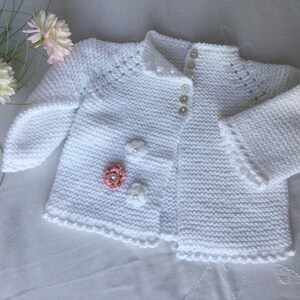 May include: White knitted baby cardigan with a button closure and decorative flowers. The cardigan has a scalloped edge.