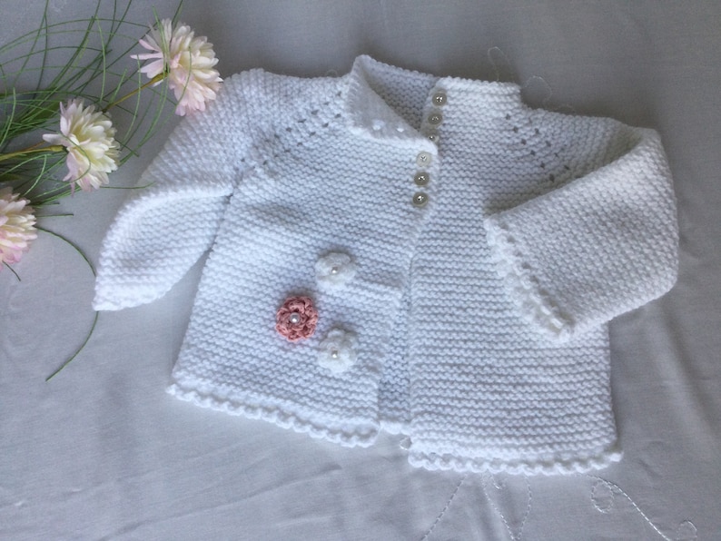 May include: White knitted baby cardigan with a button closure and decorative flowers. The cardigan has a scalloped edge.