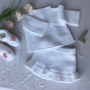 May include: A white knitted baby outfit set with a sweater and skirt. The skirt has a ruffled hem. The set is decorated with small pink flowers.