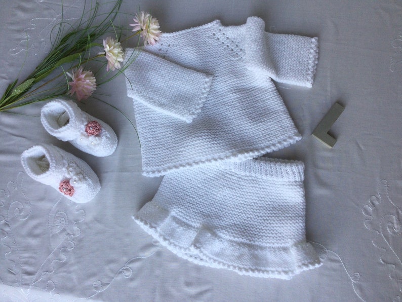 May include: A white knitted baby outfit set with a sweater and skirt. The skirt has a ruffled hem. The set is decorated with small pink flowers.