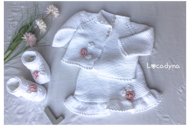 May include: White knitted baby outfit set with a cardigan, skirt, and booties. The cardigan and skirt have delicate white crocheted flowers with pink centers. The booties have pink flowers and pearl buttons.