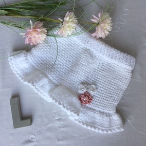 May include: White knitted baby bonnet with a ruffled edge and a pink flower embellishment.