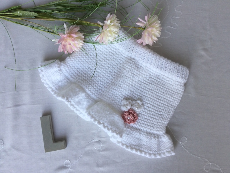 May include: White knitted baby bonnet with a ruffled edge and a pink flower embellishment.
